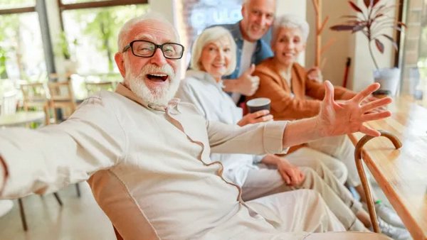 Радостный пожилой мужчина улыбается, широко раскинув руки, с тремя друзьями на заднем плане